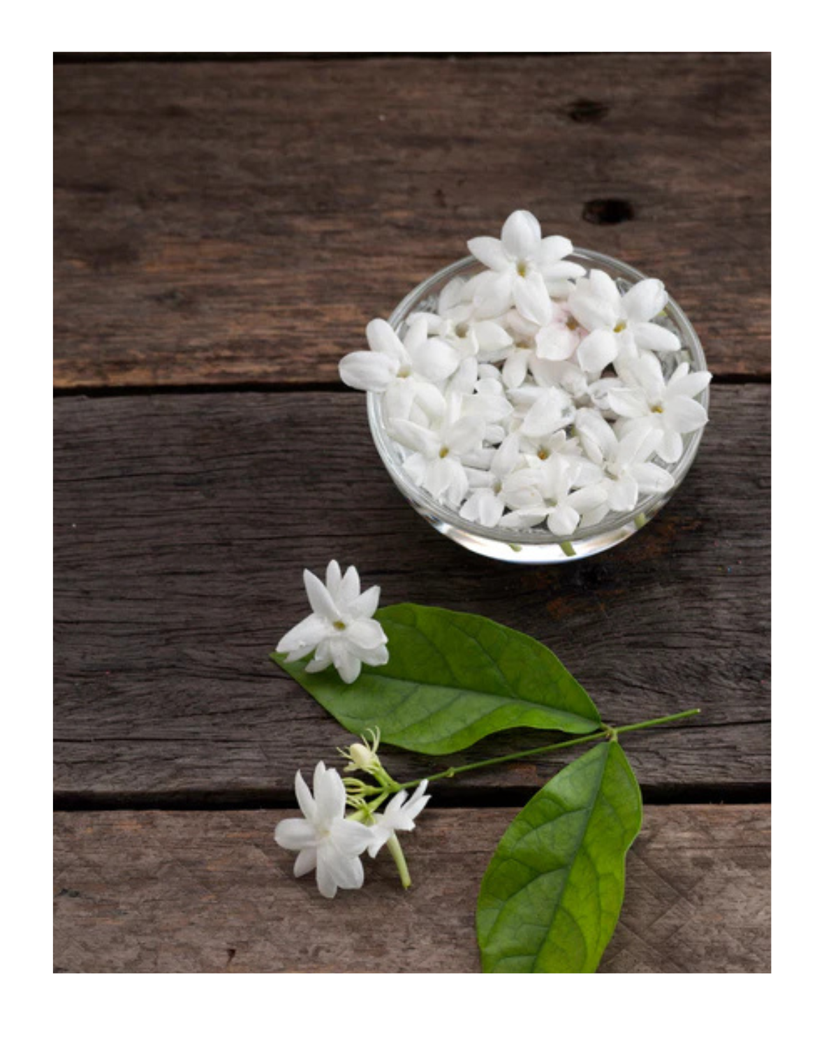 White flowers in a glass bowl on a wooden surface with green leaves.