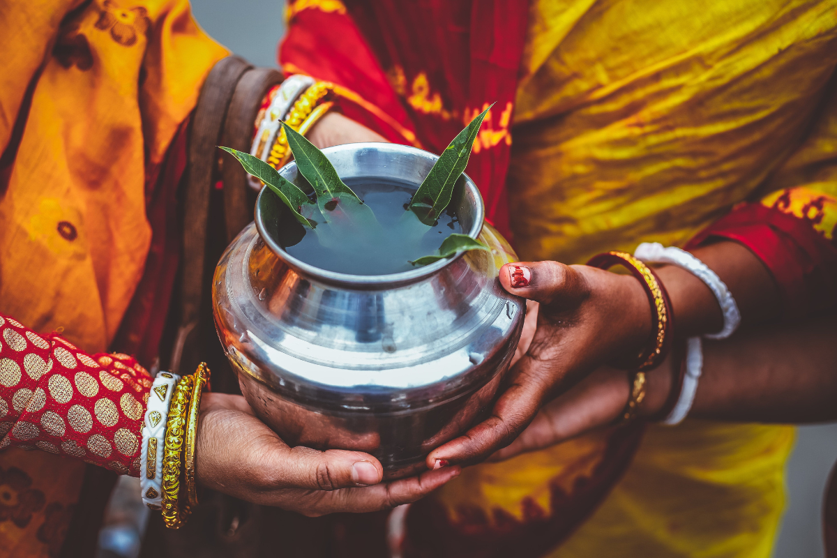 Person holding a silver pot with leaves, wearing traditional attire and jewelry.